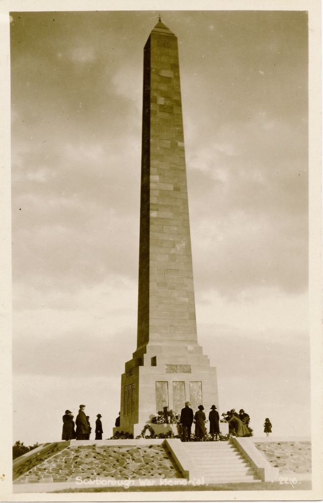 A sepia photo showing Scarborough War Memorial, a stone obelisk on top of a square pedestal on a square mound, reached by steps. Plaques can be seen on the sides of the memorial and a number of people surround the memorial, viewing the wreaths. 